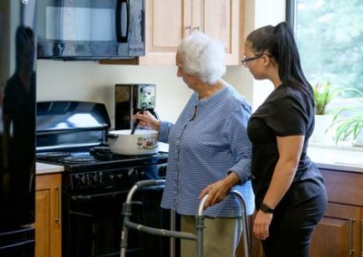 A younger woman stands beside an older with a walker while she cooks on a stove