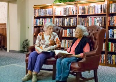 Two women sit in leather chairs in a library reading books