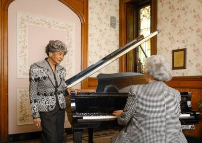 A woman stands beside a piano while another plays
